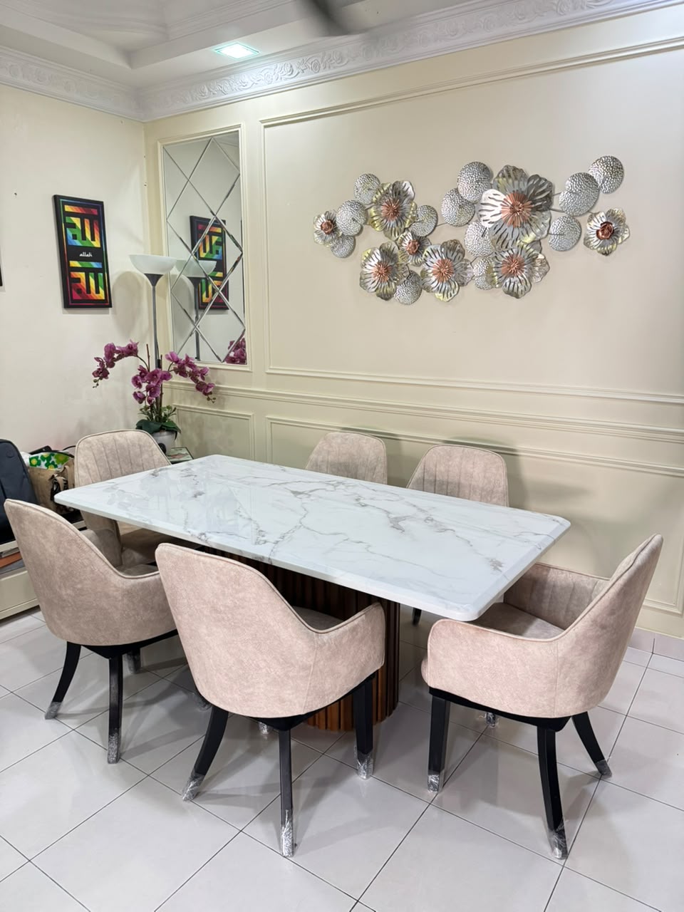 A modern dining room setup featuring a rectangular white marble-top table surrounded by six blush pink upholstered chairs with black legs. The room has cream-colored walls with decorative molding, a silver floral wall sculpture, and a tiled floor.
