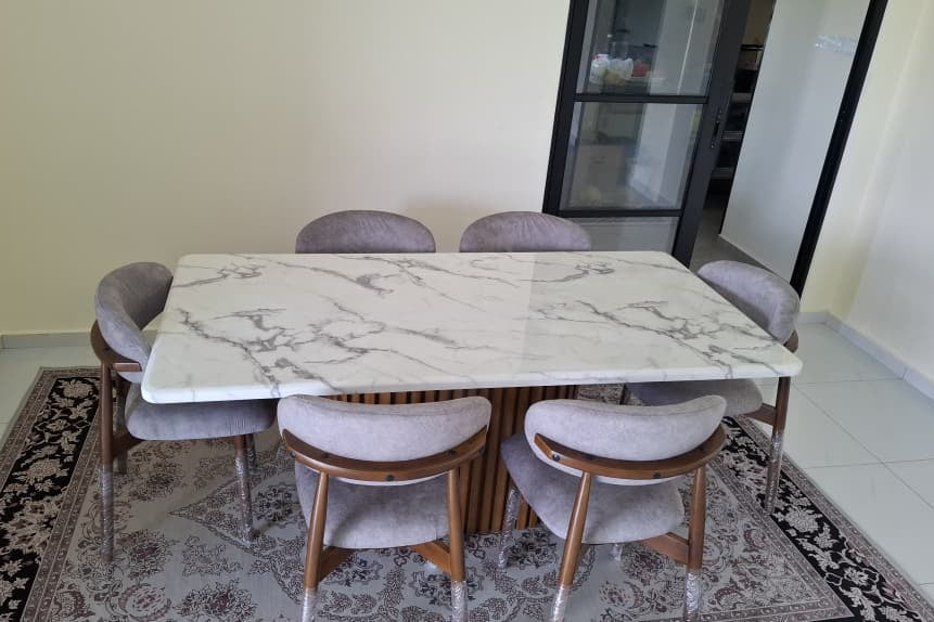 A modern dining table set featuring a white marble-patterned rectangular top with a slatted wood base, surrounded by six gray upholstered chairs with wooden frames, placed on a patterned rug in a room.
