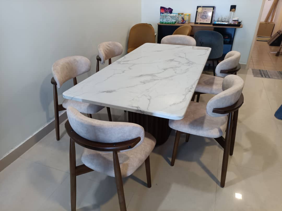 A modern dining set featuring a rectangular white marble-patterned table with a dark wood base, surrounded by six upholstered chairs with beige fabric and dark wood frames, set in a home interior.