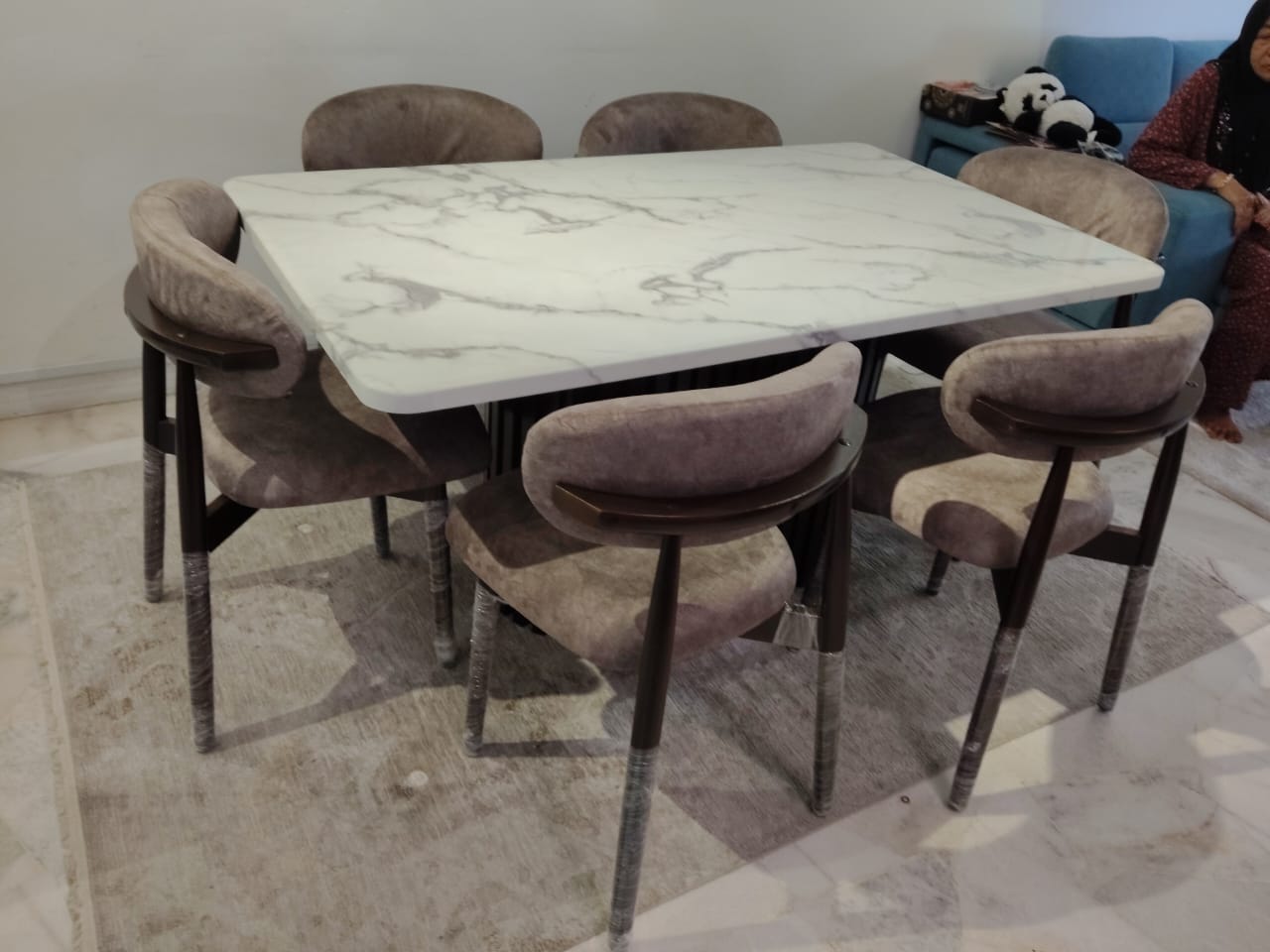 A modern 7-piece dining set featuring a square white marble-patterned table and six upholstered velvet chairs in a dusty mauve-gray color, set on a light rug in an indoor room.