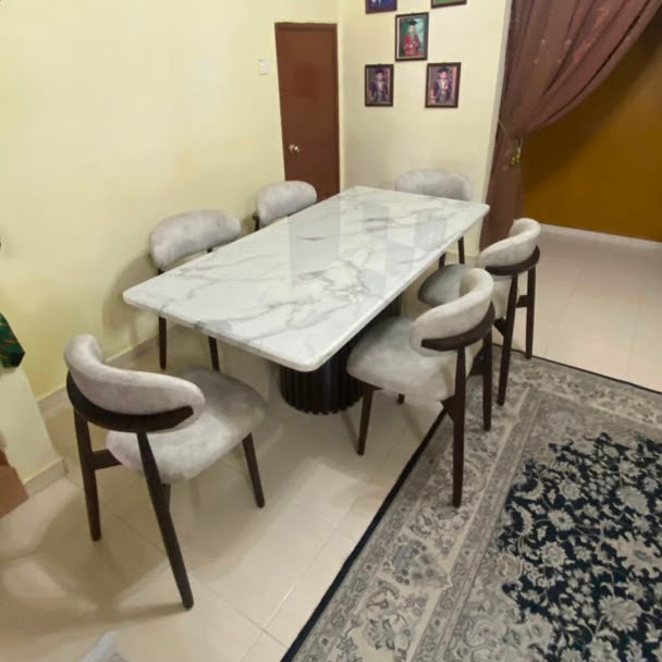 A modern dining room setup featuring a rectangular white marble-patterned dining table surrounded by six upholstered dining chairs with dark wood legs, set on a patterned area rug in a home interior.