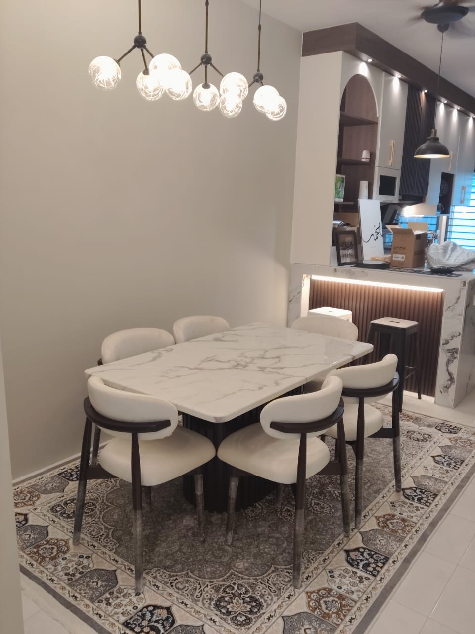 A modern dining room setup featuring a rectangular white marble-top table surrounded by six upholstered dining chairs with dark wood frames, set on a patterned area rug. The room has warm neutral walls, a multi-light spherical pendant chandelier, and a kitchen area in the background.