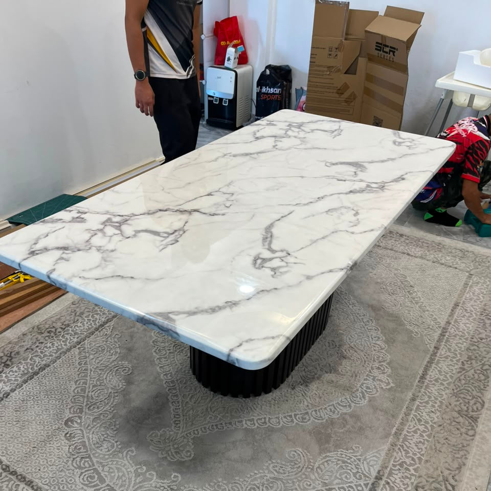 A rectangular coffee table with a white marble-patterned top and a black ribbed pedestal base, photographed in a showroom or warehouse setting on a large patterned rug. A person is partially visible at the upper left, and cardboard boxes and store items are in the background.