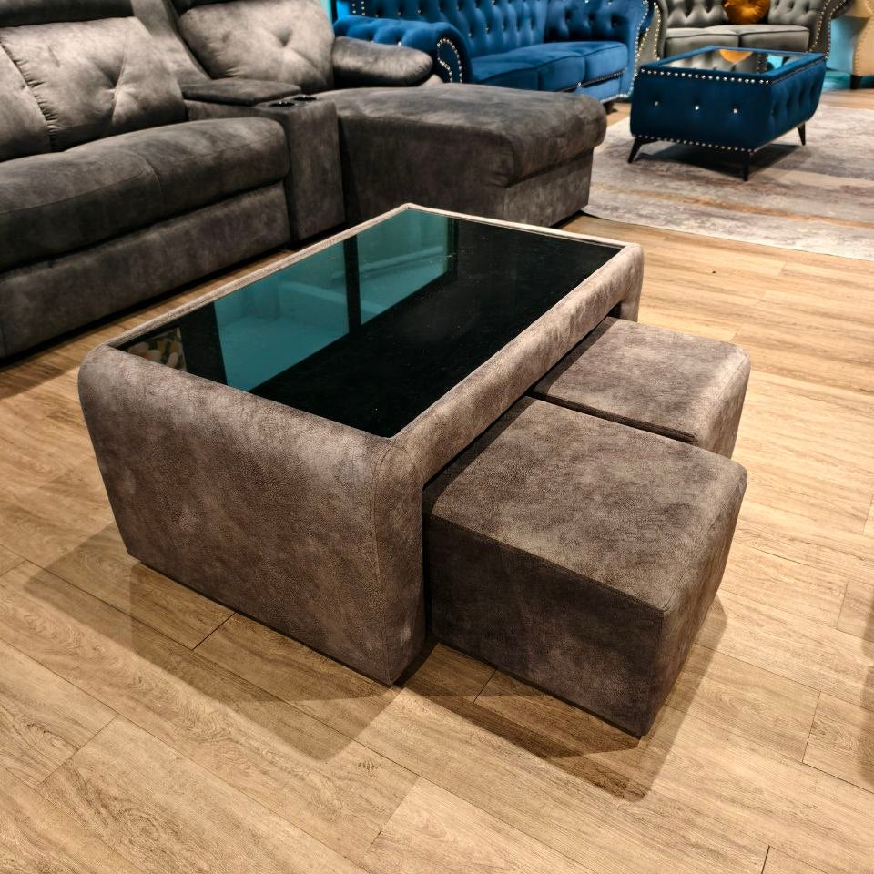 A modern coffee table with a rectangular black glass top and three matching square ottomans in a dark taupe-brown textured fabric, set on a light wood floor in a furniture showroom.